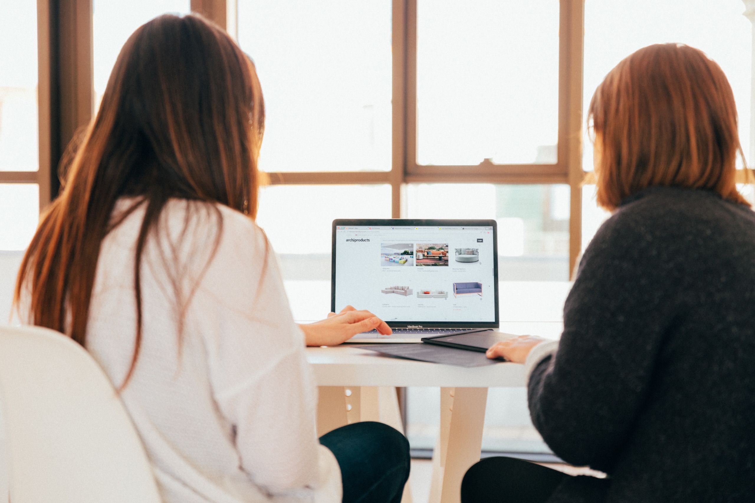 Zwei als Frauen* gelesene Personen sitzen gemeinsam vor einem Laptop vor einer großen Fensterfront.